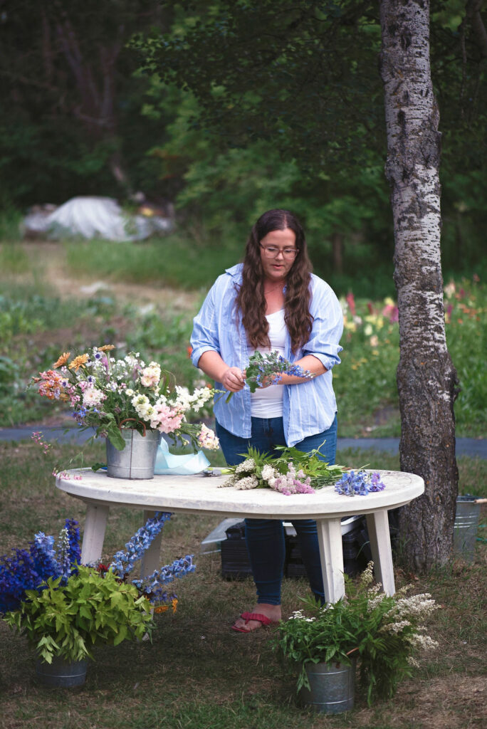 woman in blue arranging cut flowers from the garden on a table outside in front of the garden